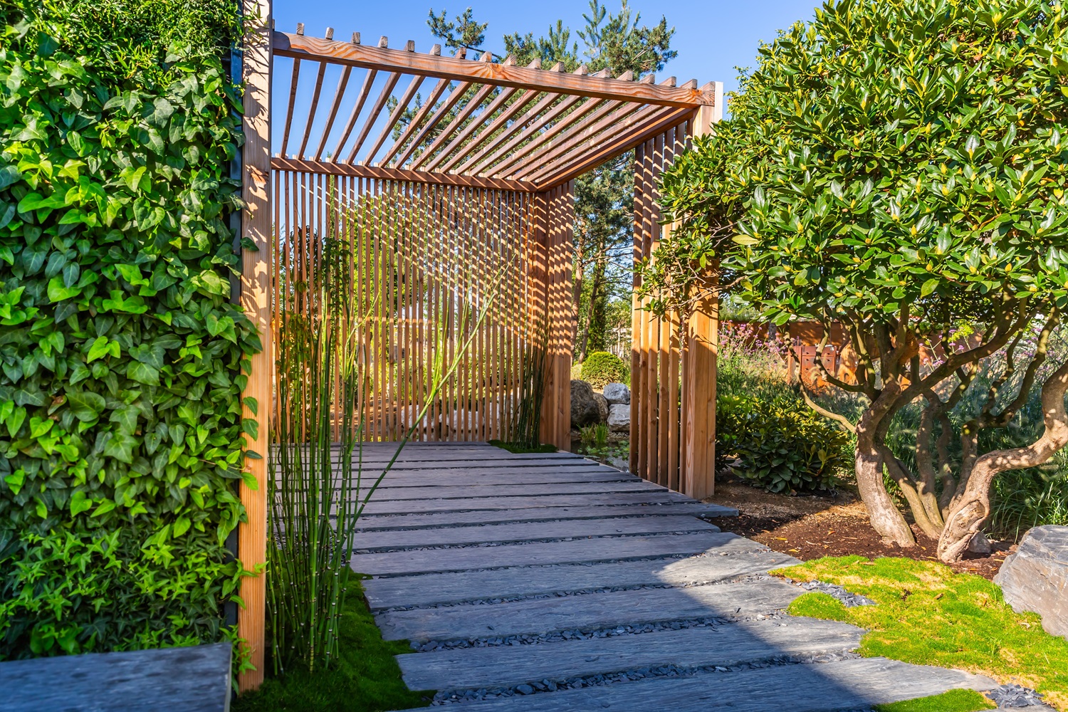 A wooden pergola over a stone path surrounded by greenery and trees