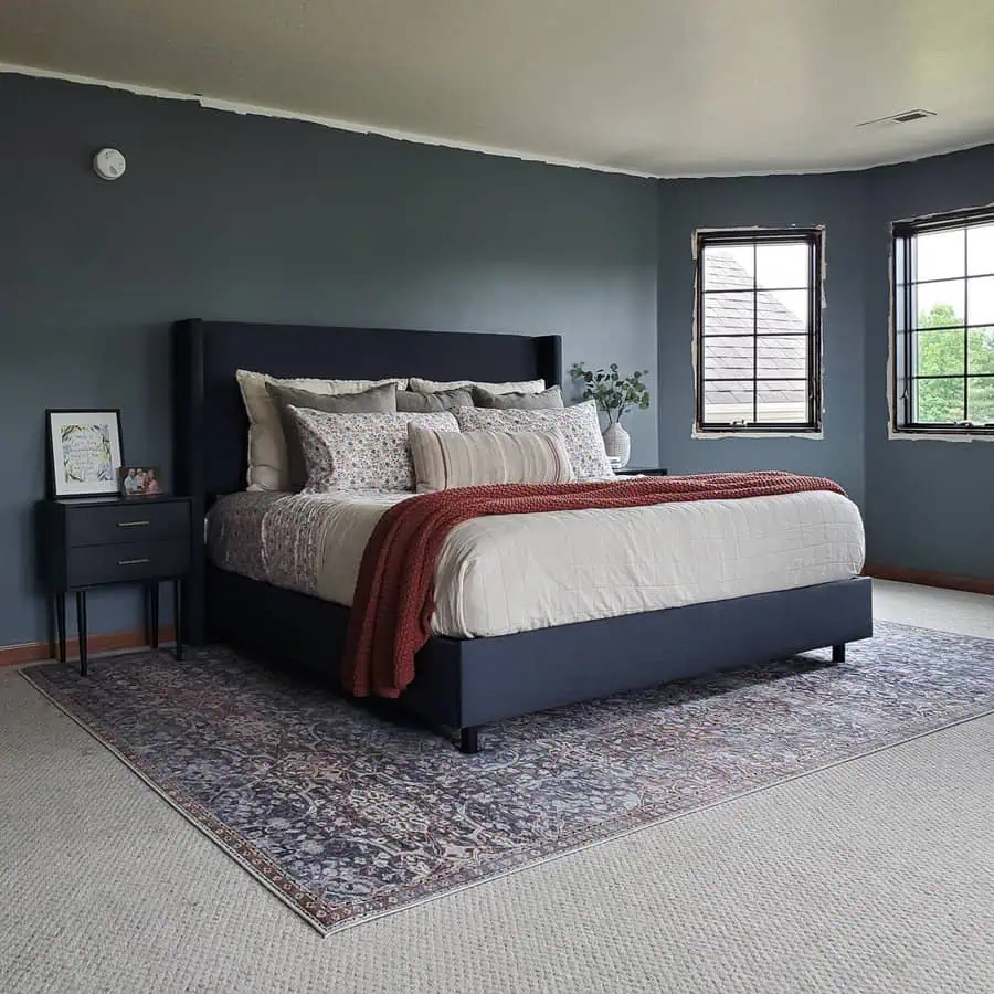 Cozy bedroom with dark blue walls, upholstered bed, patterned rug, and layered bedding with a red throw blanket.