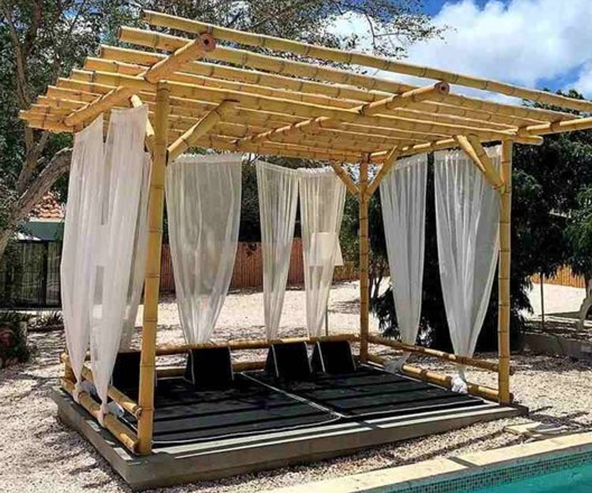 A bamboo cabana with white curtains and black lounge chairs by a swimming pool