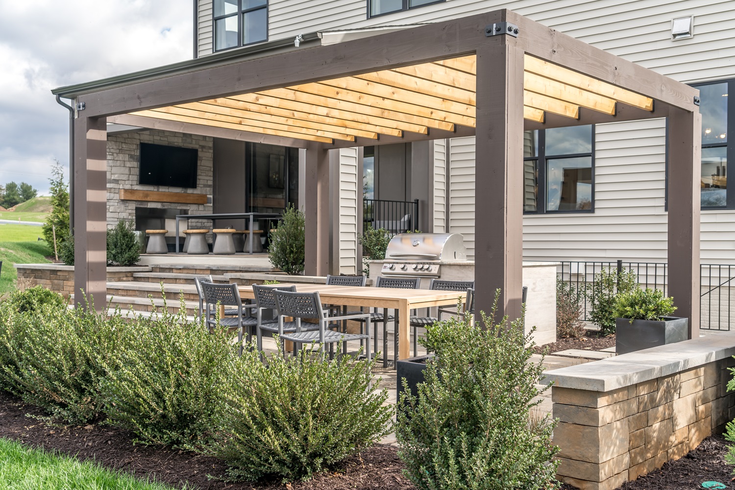 Outdoor patio with a pergola, dining table, chairs, and a grill surrounded by greenery