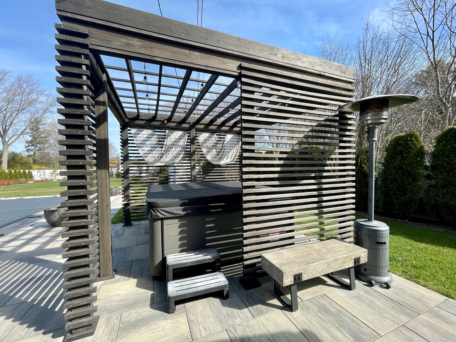 Outdoor pergola with hot tub, steps, bench, and patio heater on a sunny day