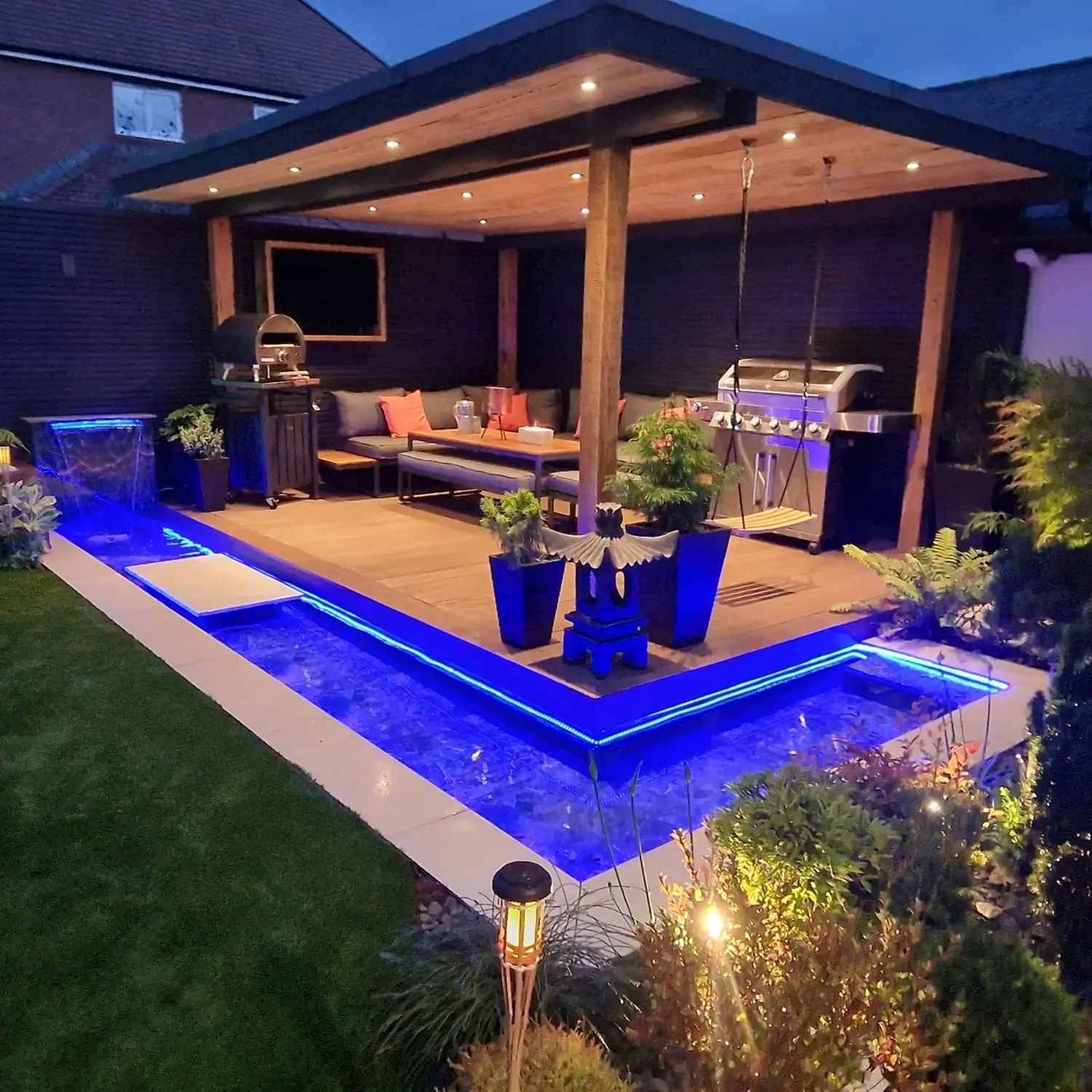 Modern outdoor patio featuring a blue LED-lit pool and a covered seating area, illuminated at night