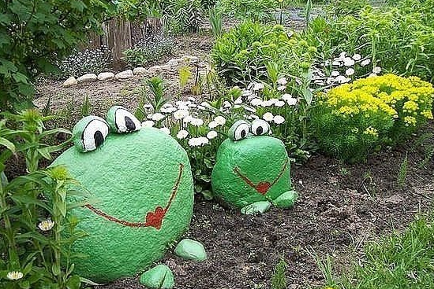 Two frog-shaped garden rocks with painted faces amidst flowers and greenery