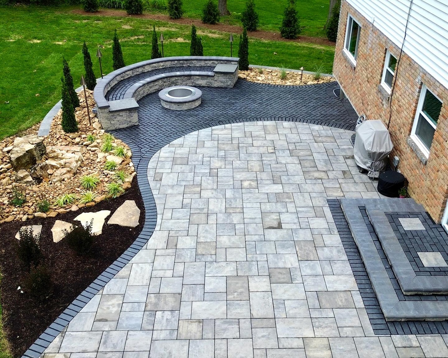 A backyard patio with gray interlocking pavers, curved seating wall, a fire pit, a barbecue grill, and landscaped with small trees and rocks near a brick house.