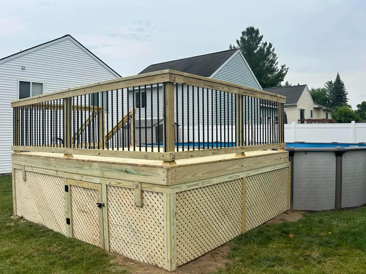 Wooden deck with storage with black railings around an above-ground pool in a suburban backyard.