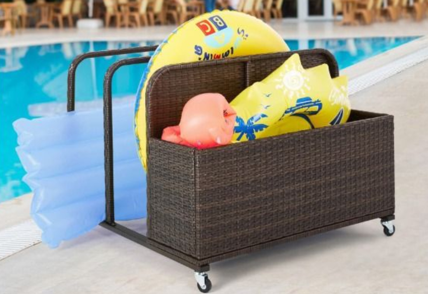 A wicker storage cart with wheels is filled with colorful pool inflatables, including a yellow float ring and a blue lounger, positioned beside a swimming pool.