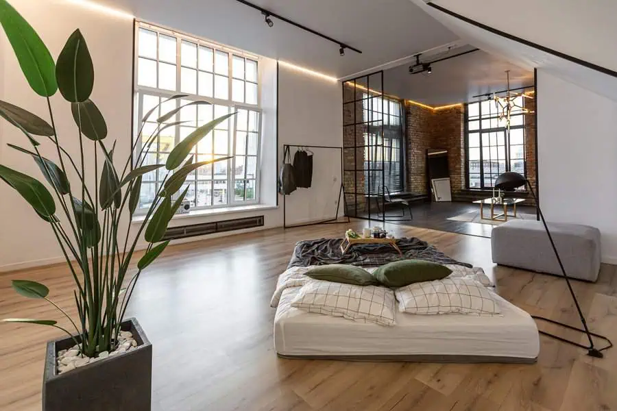 Loft bedroom with large windows, minimalistic decor, and a low bed on wooden flooring.