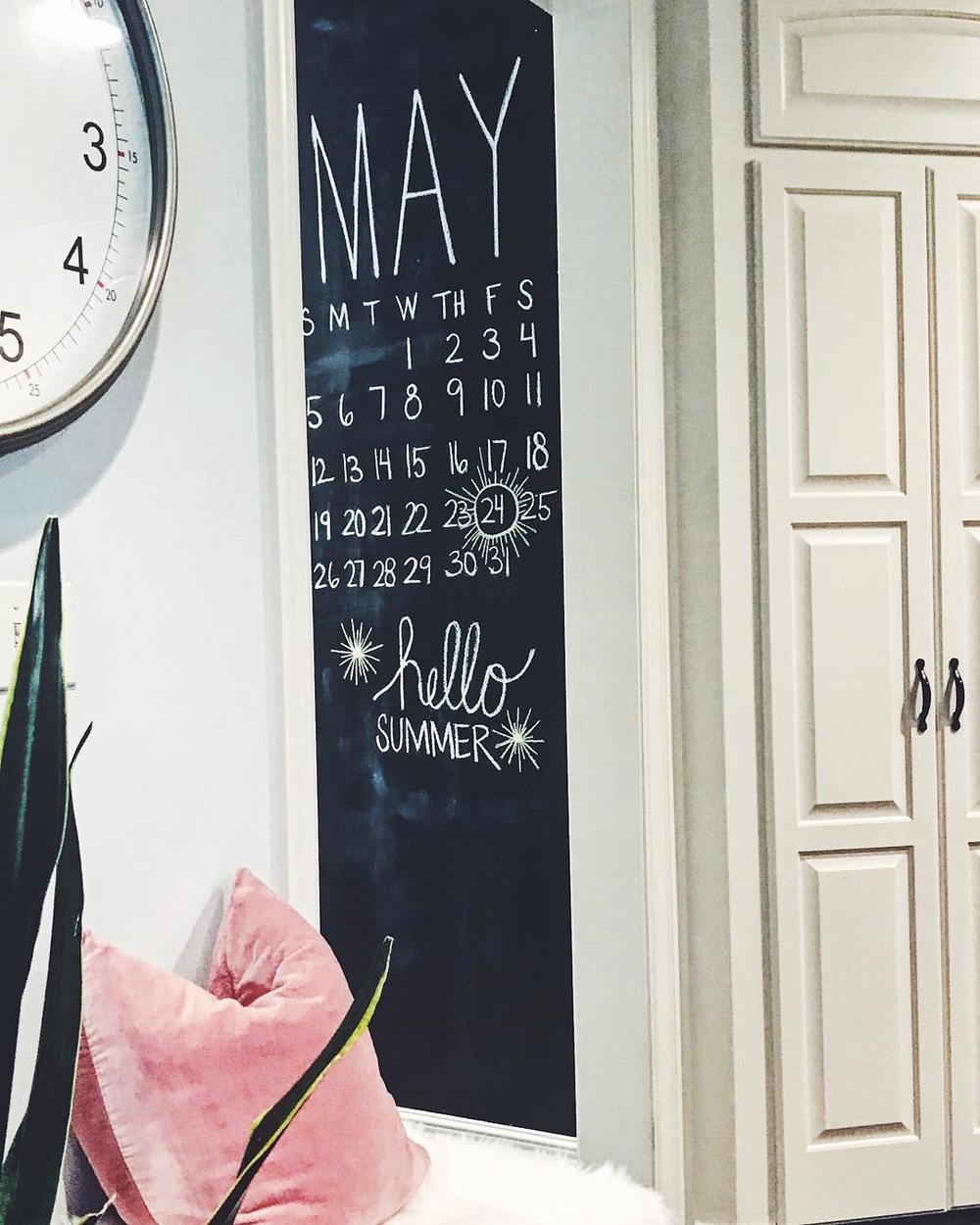 Blackboard calendar for May with "hello SUMMER" written below and tropical decor nearby.