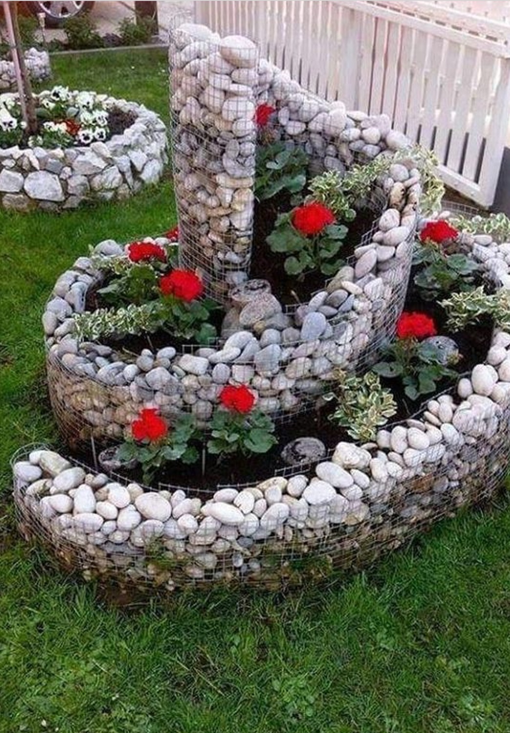 A spiral garden bed made of rocks and wire, filled with vibrant red flowers and lush green plants