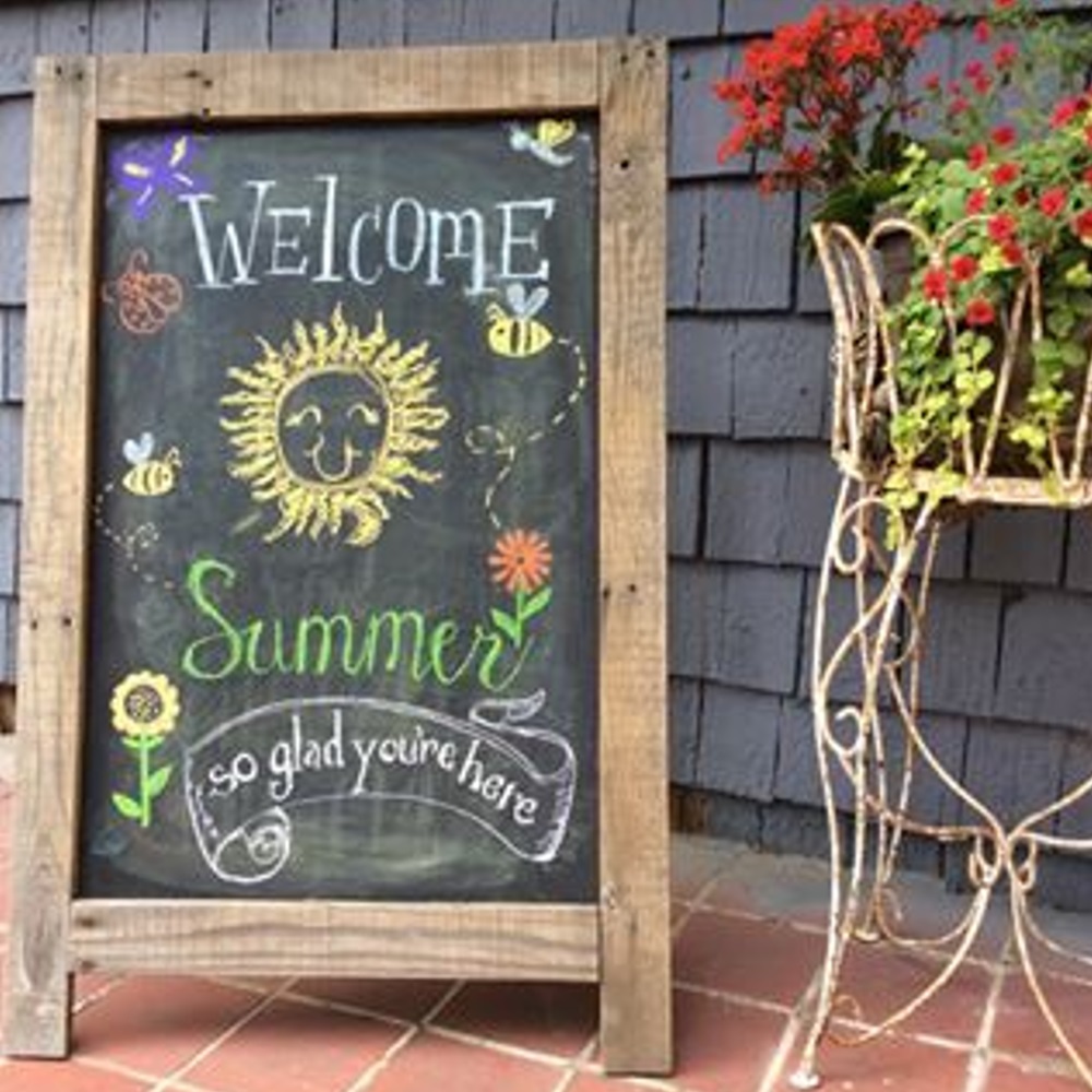 Decorative chalkboard with "Welcome Summer" message next to a plant stand with flowers.