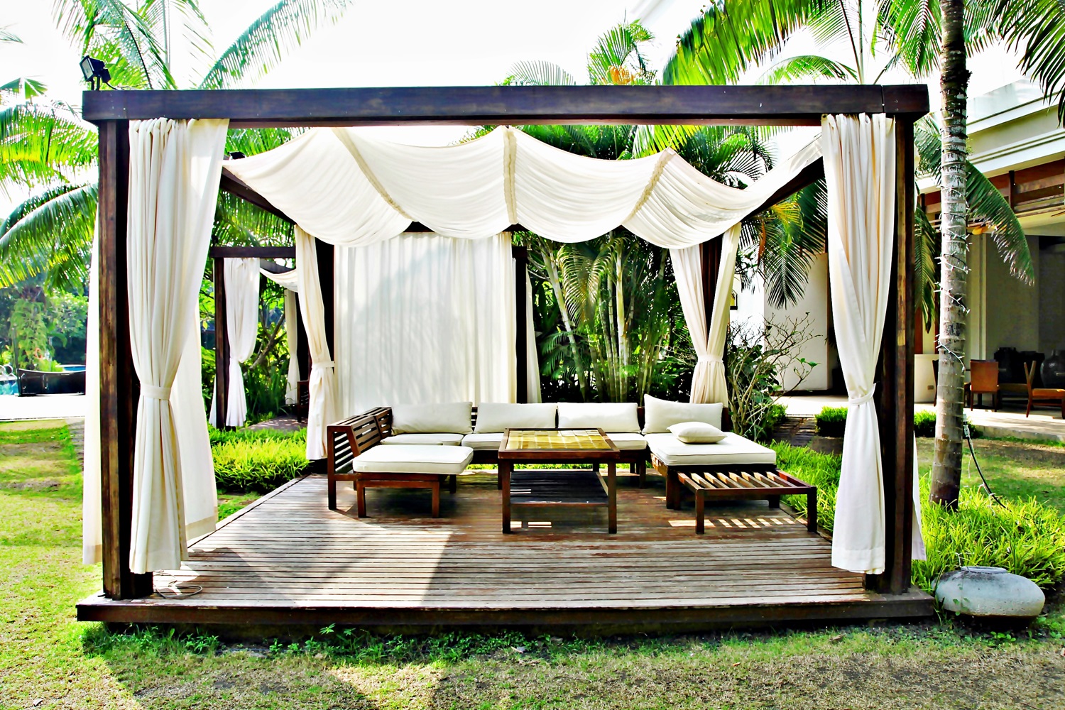 A shaded outdoor lounge with white curtains, sofas, and wooden furniture on a deck