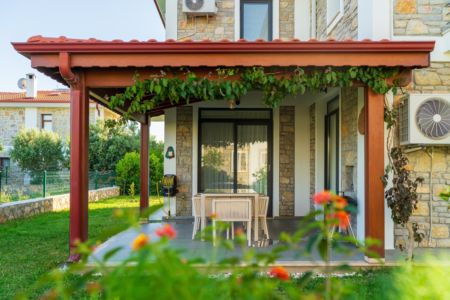 Covered patio with dining table and chairs outside a stone house with greenery