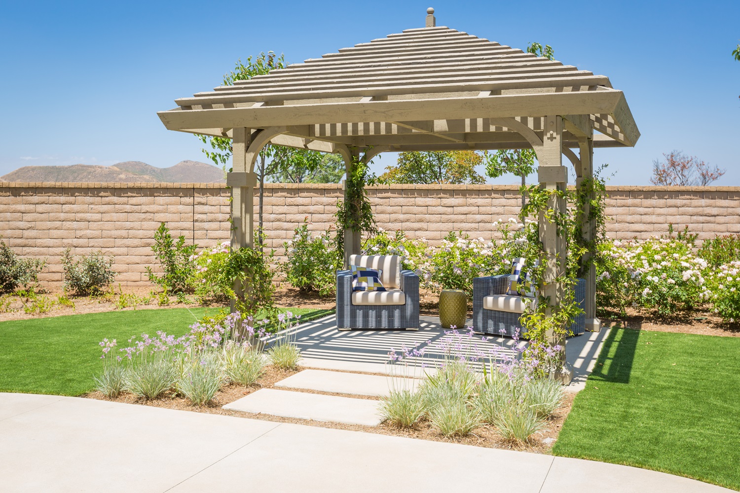 A backyard patio featuring a pergola, two chairs, flowers, and a stone-paved walkway