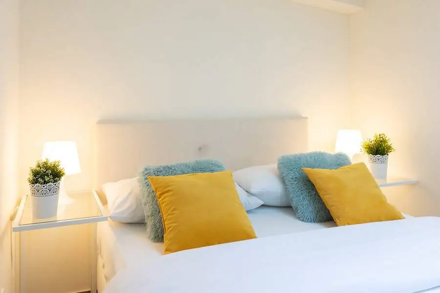 Cozy bedroom with a white bed, yellow and teal accent pillows, and bedside tables with lamps and plants.