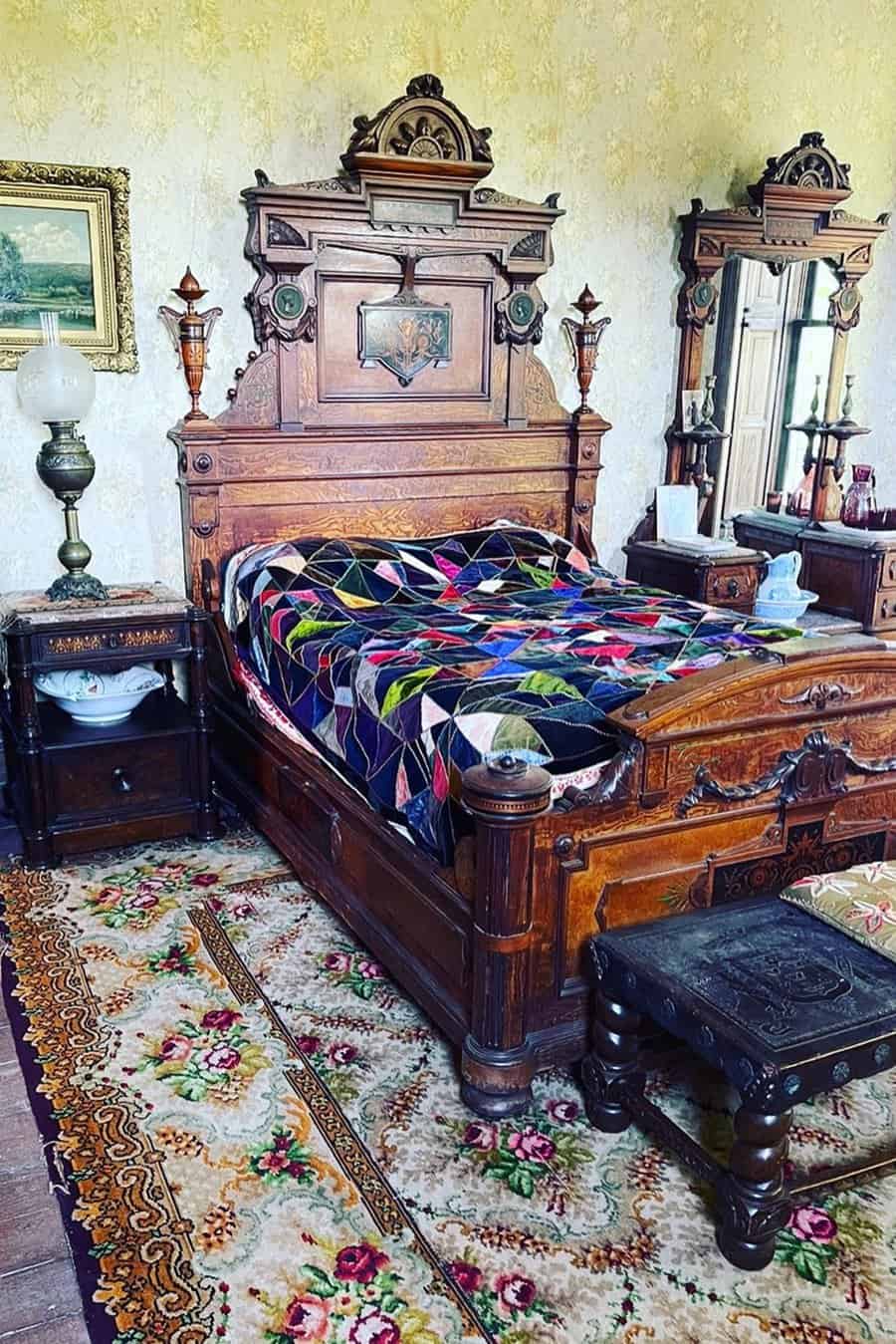 Vintage bedroom with ornate wooden bed, colorful quilt, antique furniture, oil lamp, and floral-patterned rug