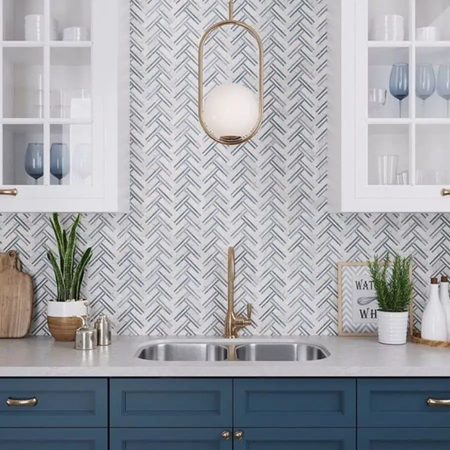 A kitchen with blue lower cabinets, a herringbone tile backsplash, and a brass faucet under a modern pendant light.