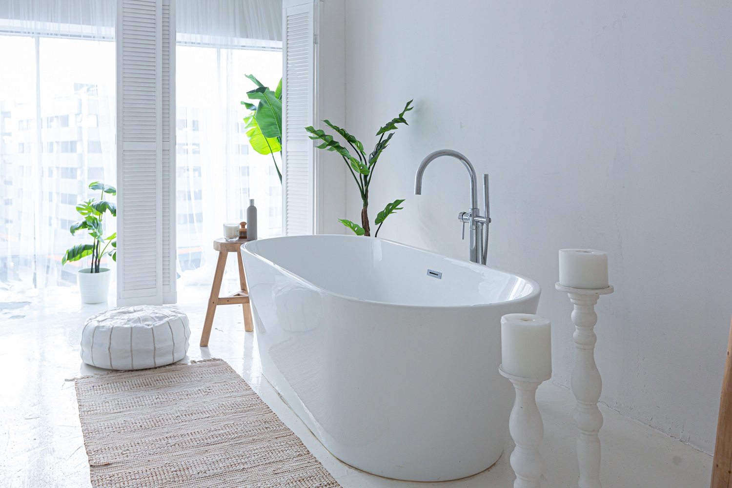 Minimalist white bathroom with a freestanding tub, plants, stool, candles, and a pouf