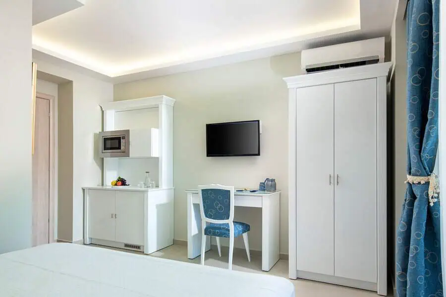 Modern hotel room featuring white furniture, wall-mounted TV, microwave, and a blue-accented chair