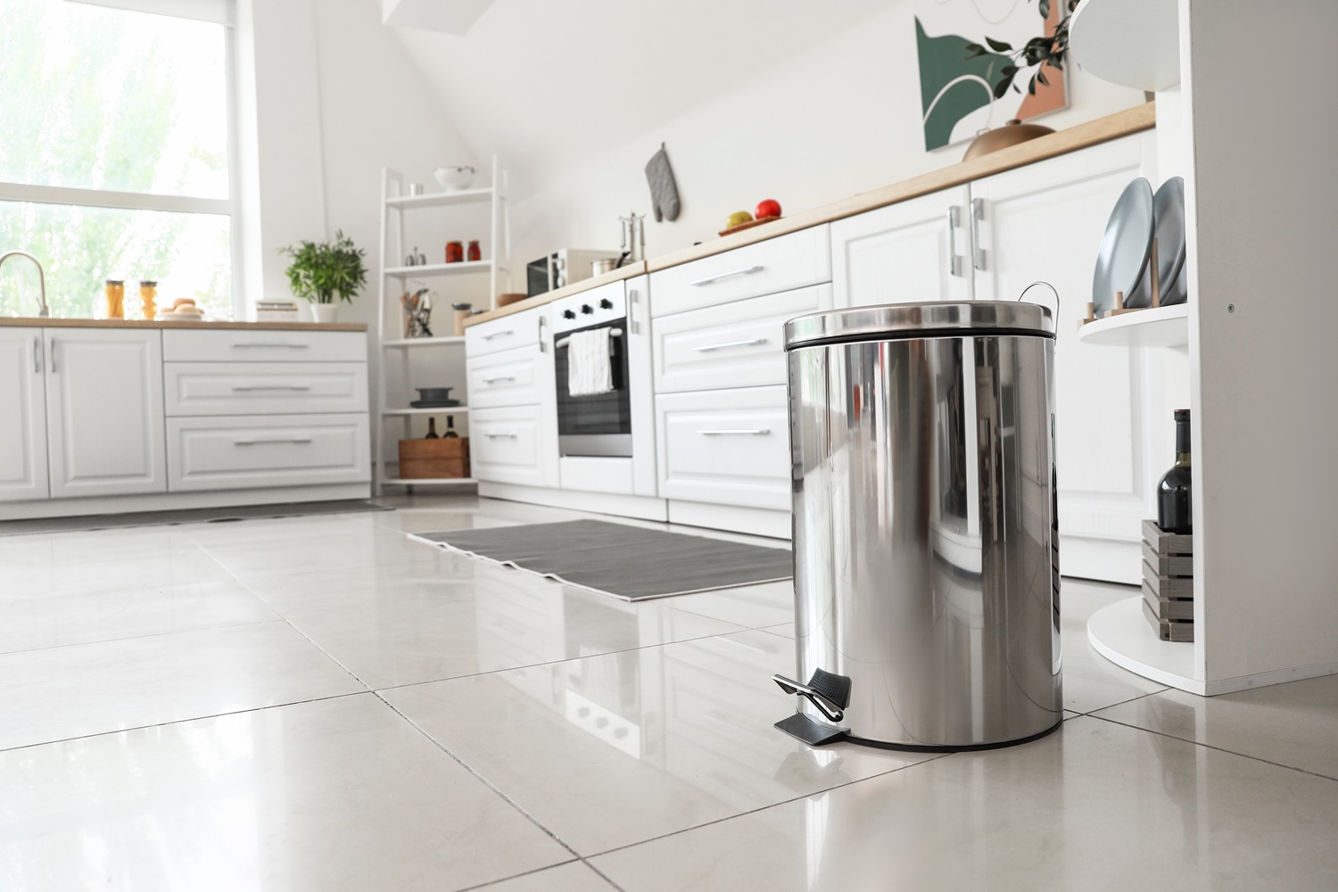 Modern white kitchen with a stainless steel trash bin on a tiled floor