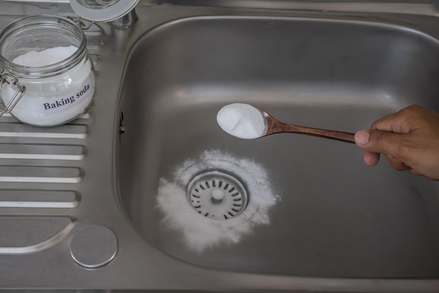 Hand pouring baking soda from a spoon into a sink