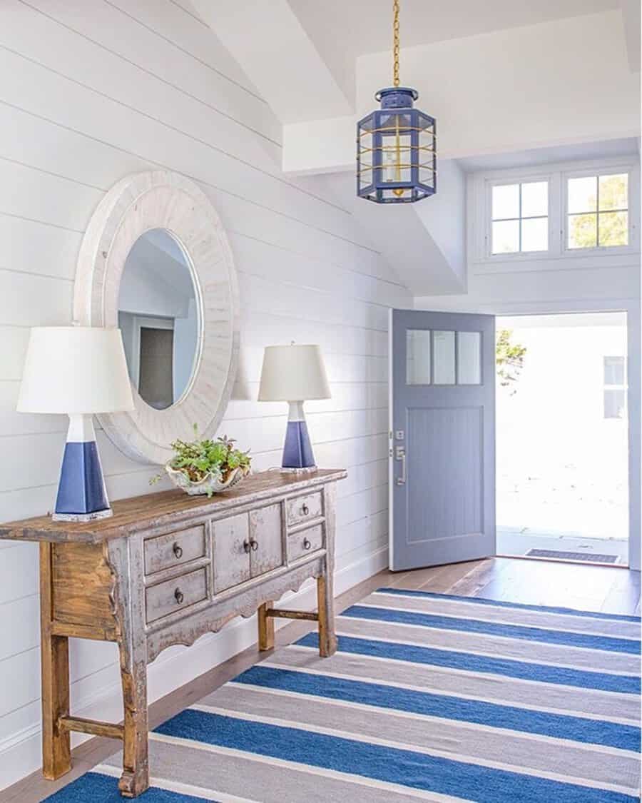 Bright entryway with a rustic wooden console table, round mirror, blue and white lamps, and a striped blue rug