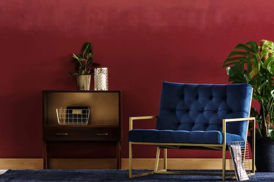 Stylish living room with a blue armchair, golden frame, wooden side table holding plants and a decorative basket, all set against a rich red wall and dark rug.