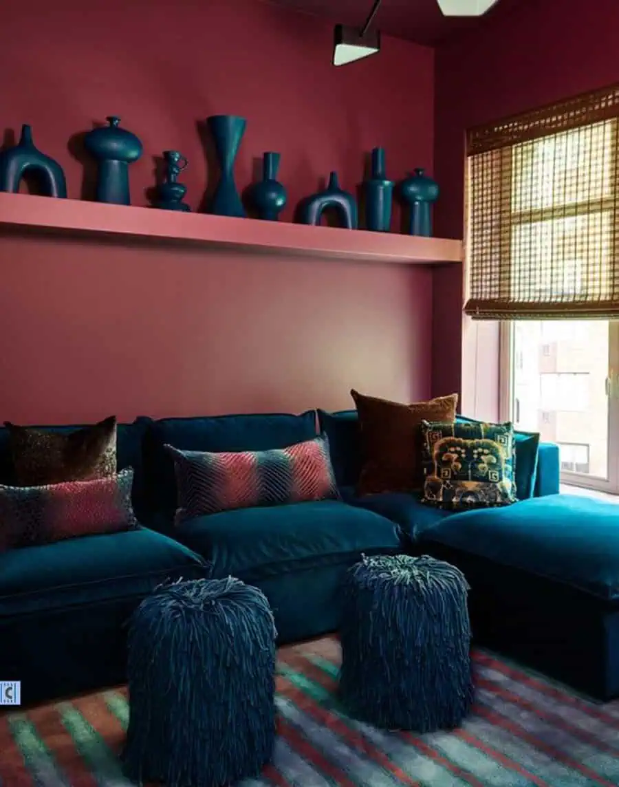 A living room has bold burgundy walls and a ceiling, a teal sectional sofa with textured pillows, blue fuzzy stools, and artistic vases on a shelf above the sofa.