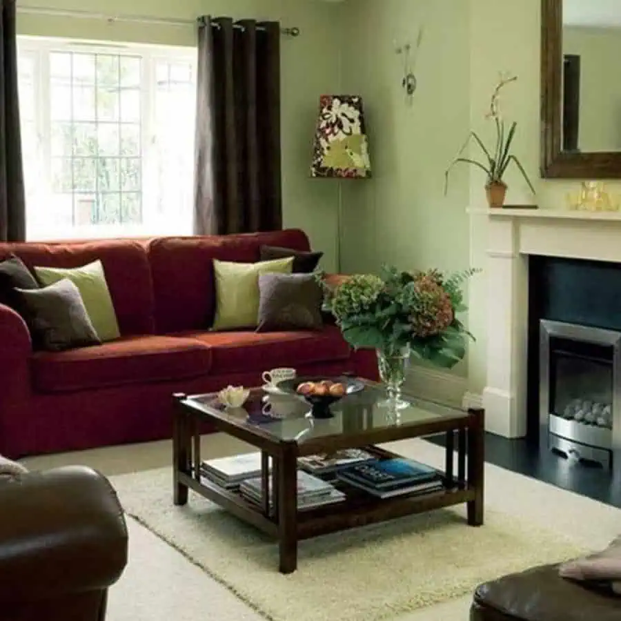 A cozy living room with a red sofa adorned with green and patterned cushions, a wooden coffee table, a fireplace, a mirror above it, and a bouquet of flowers.