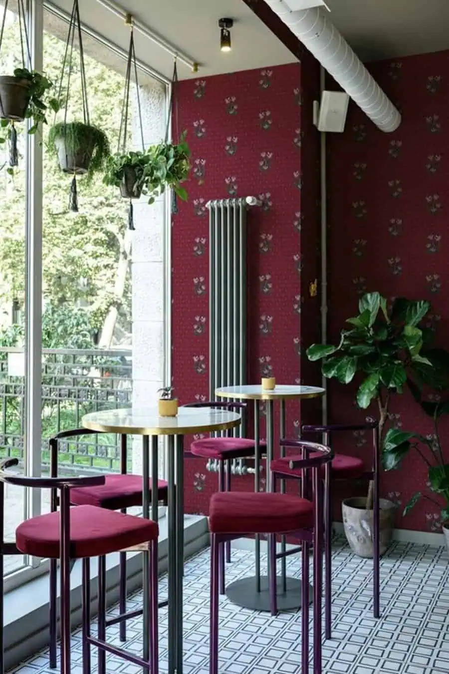 Cozy café corner with burgundy walls, high tables, velvet chairs, and hanging plants by large windows.