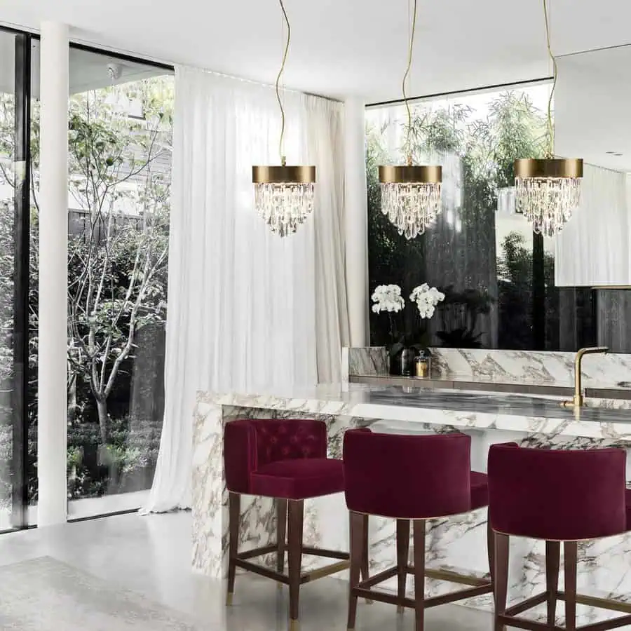 Elegant kitchen with a marble island, burgundy velvet barstools, and crystal chandeliers, overlooking a lush garden.