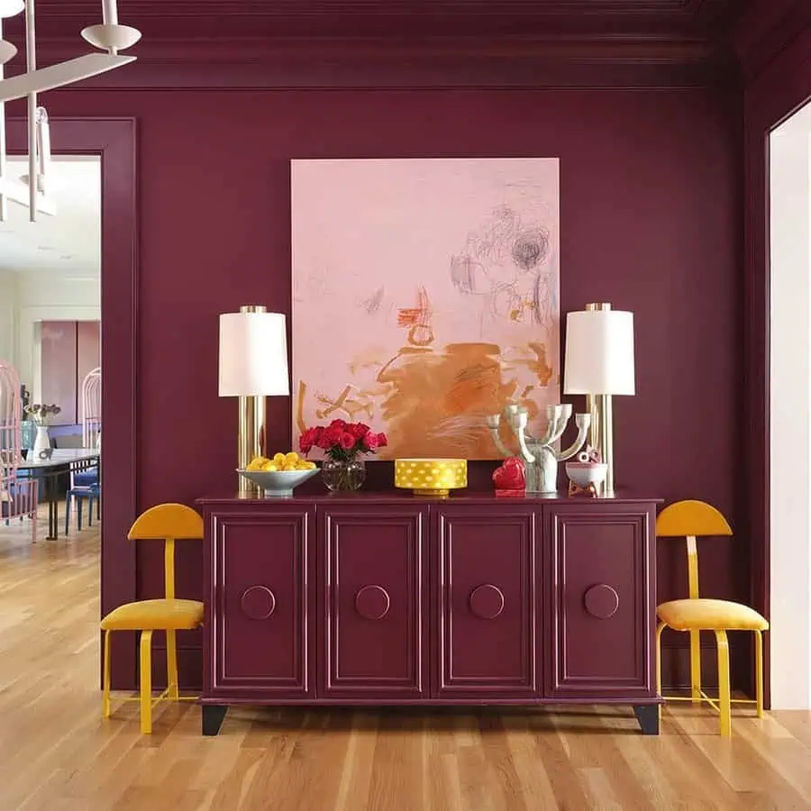 A modern room with burgundy walls, a matching sideboard, yellow chairs, abstract artwork, table lamps, decorative items, and wooden flooring.