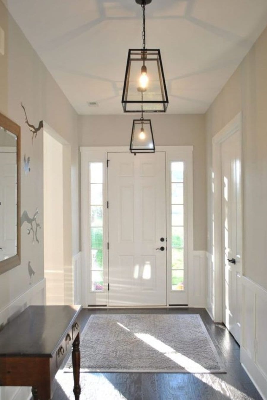 Bright entryway with glass-paneled front door, two hanging light fixtures, wall mirror, and a wooden table with drawers