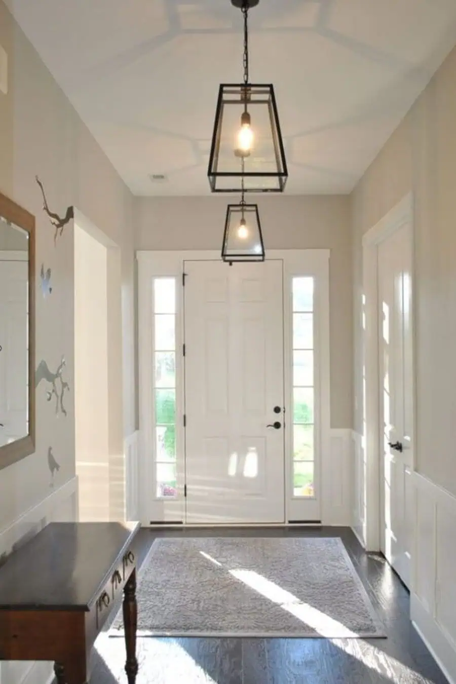 Bright entryway with glass-paneled front door, two hanging light fixtures, wall mirror, and a wooden table with drawers