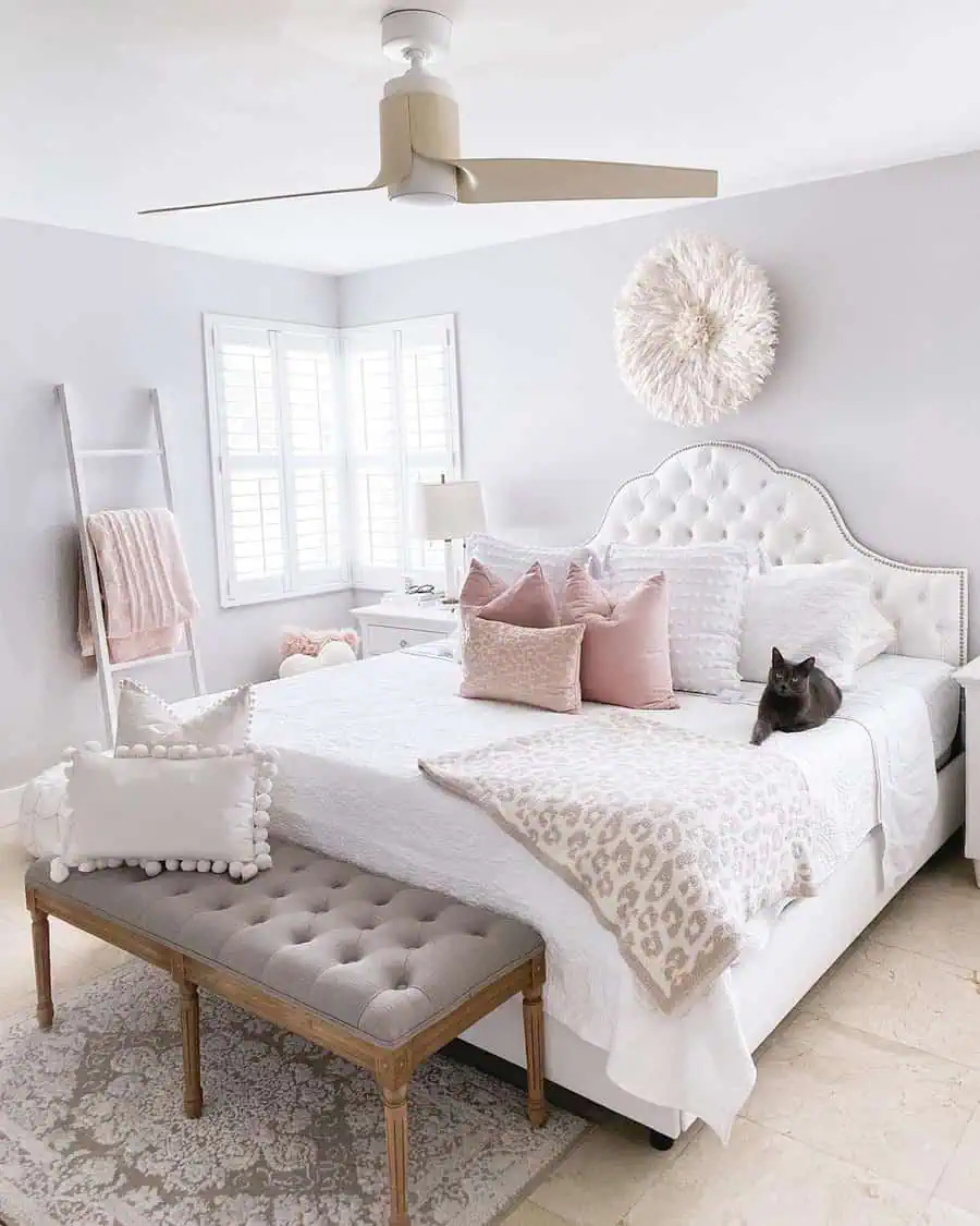 Bright and airy bedroom with white and blush decor, a tufted headboard, and a gray cat lounging on the bed.