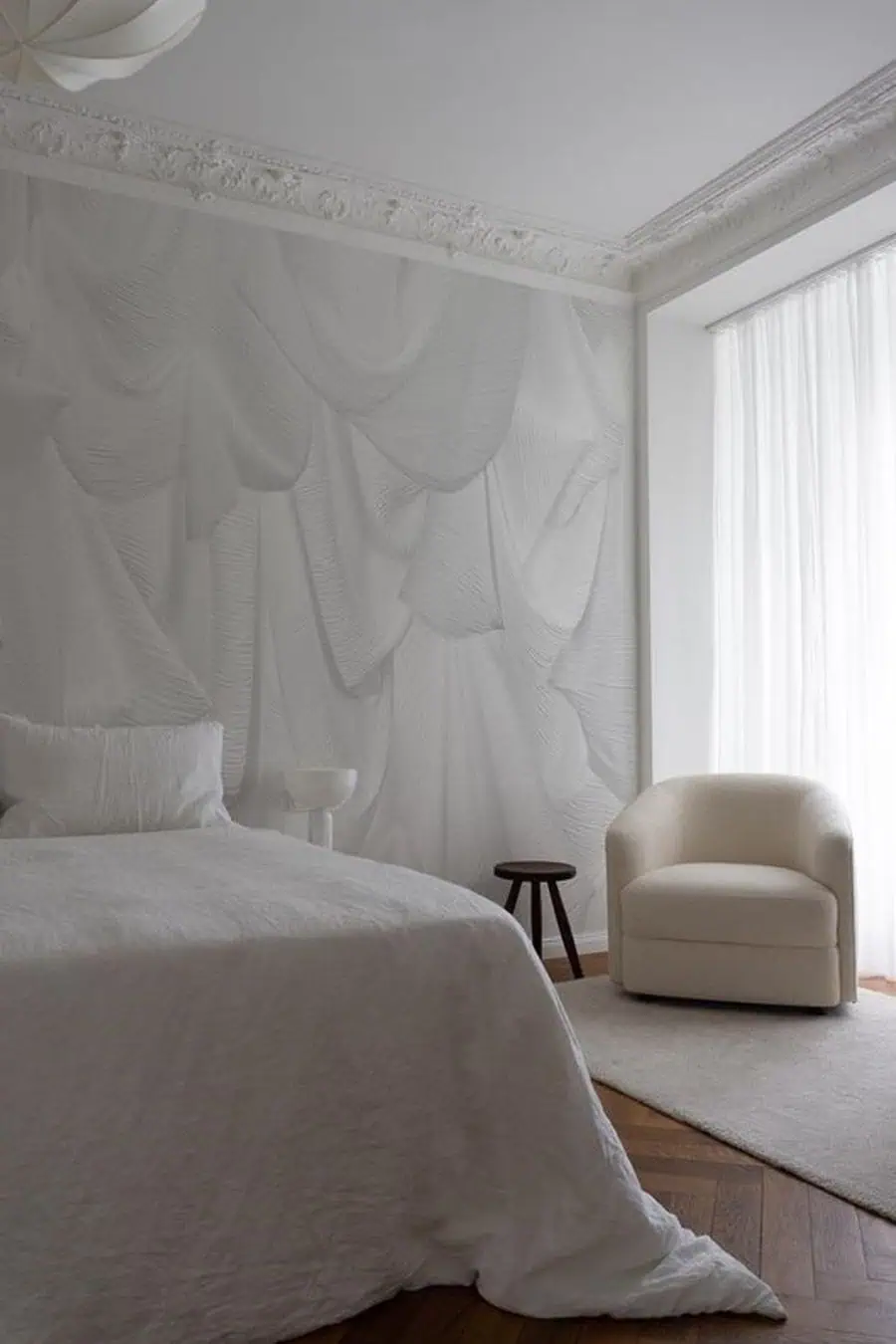 Elegant bedroom with textured walls, a large white bed, and a cozy armchair near a bright window.