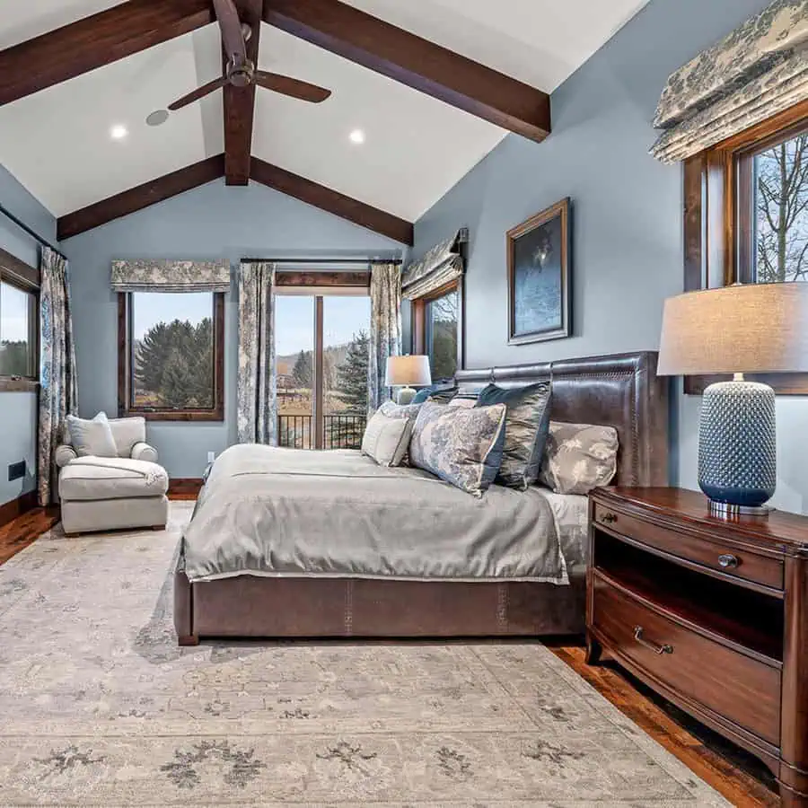 Spacious bedroom with wooden beams, large bed, armchair, blue walls, and forest view windows