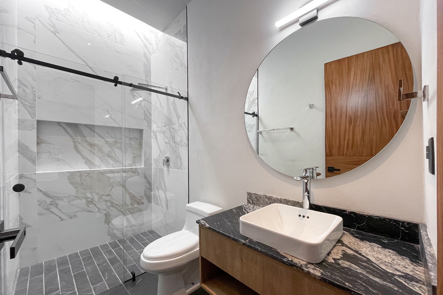 Modern bathroom with round mirror, glass shower, and black marble countertop sink