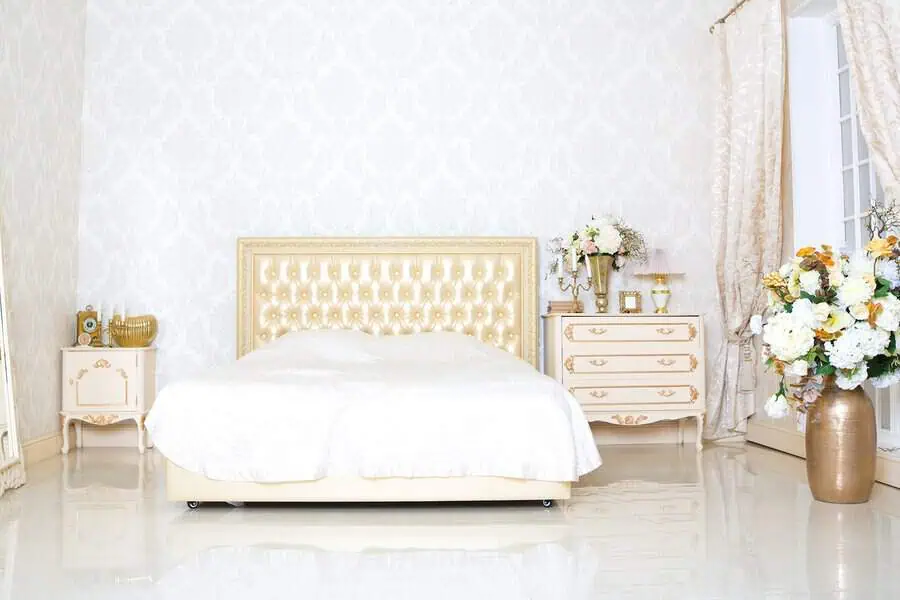 Elegant bedroom with a cream-colored palette featuring a plush, tufted bed, two ornate nightstands with decorative items, and lush floral arrangements.