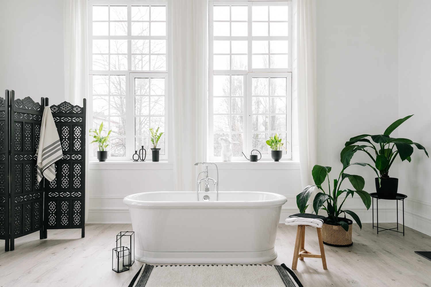 White bathroom with a bathtub, plants, large windows, and a decorative screen