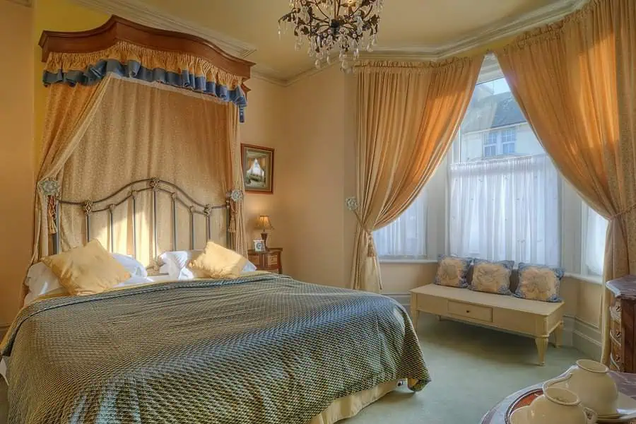 Elegant bedroom featuring a canopy bed, chandelier, large windows with draped curtains, and a bench beneath the window