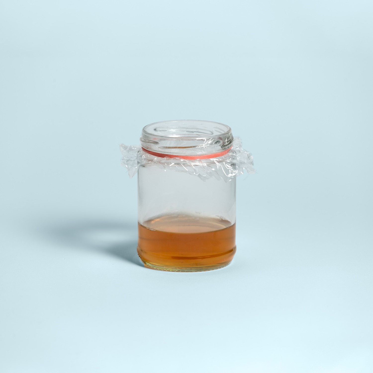 Glass jar with a small amount of amber liquid, covered with plastic wrap that serves as a Fly Trap