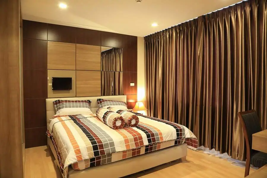 Modern bedroom with a neatly made bed, mirrored wall, wooden floor, and brown curtains.