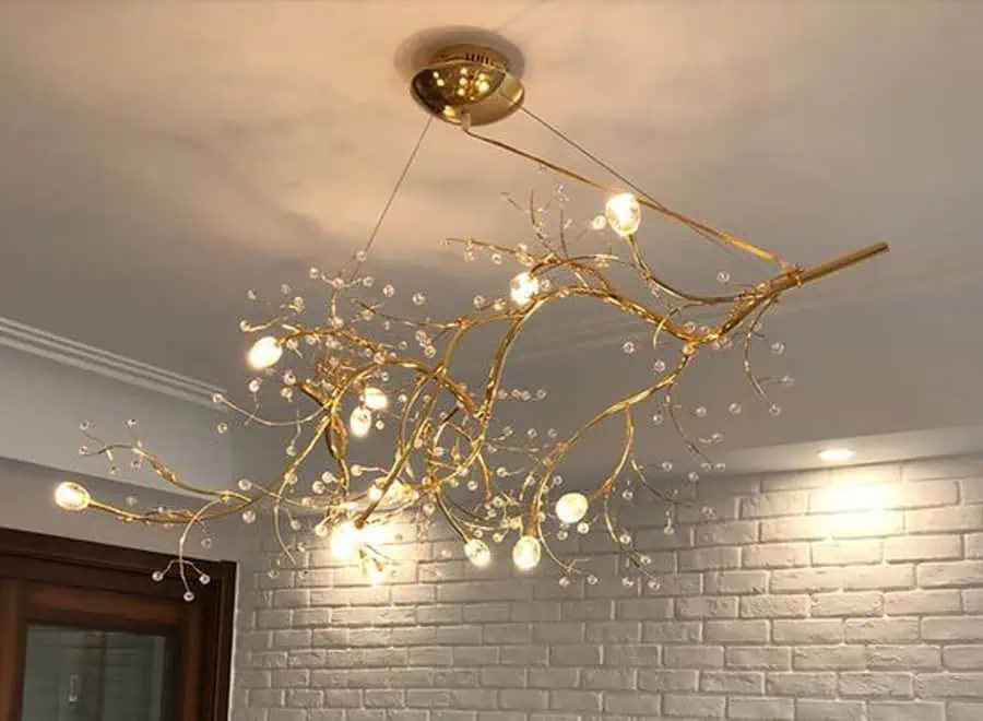 A golden, branch-like chandelier with circular lights hangs from a ceiling, illuminating a white brick wall