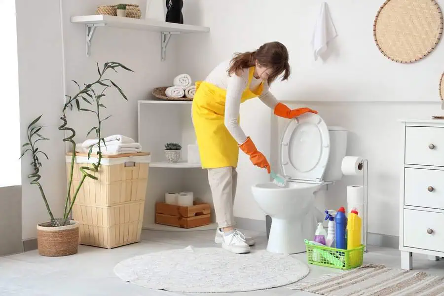 Cleaning a toilet in a bathroom with various cleaning supplies in a basket nearby