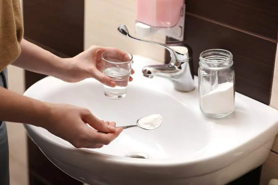 Individual holding a glass and spoon with baking soda near a sink