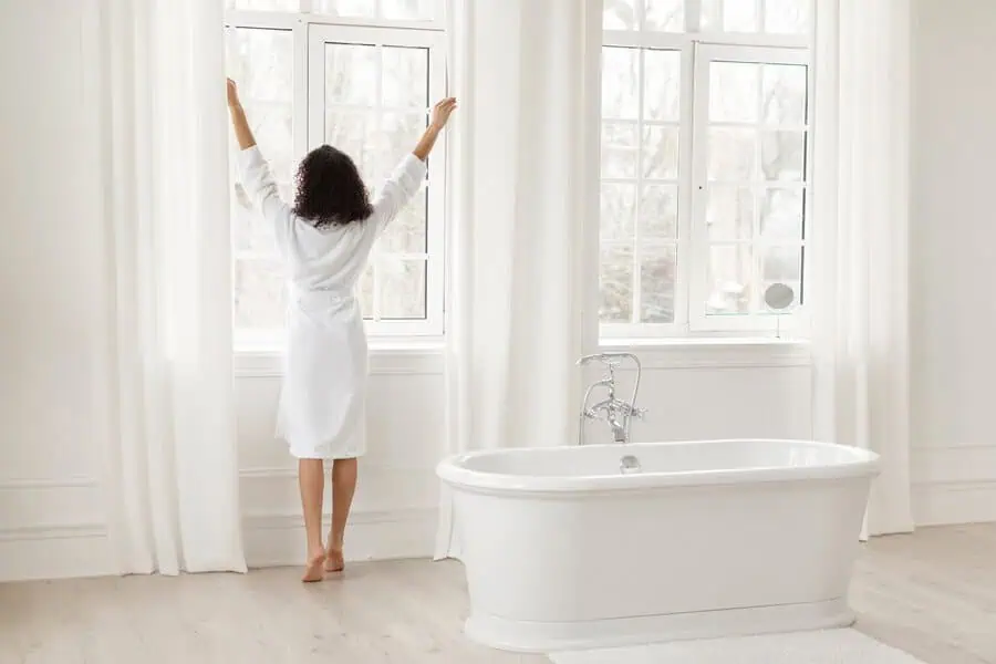 Individual in bathrobe stretching by a window in a bright, white bathroom with a tub