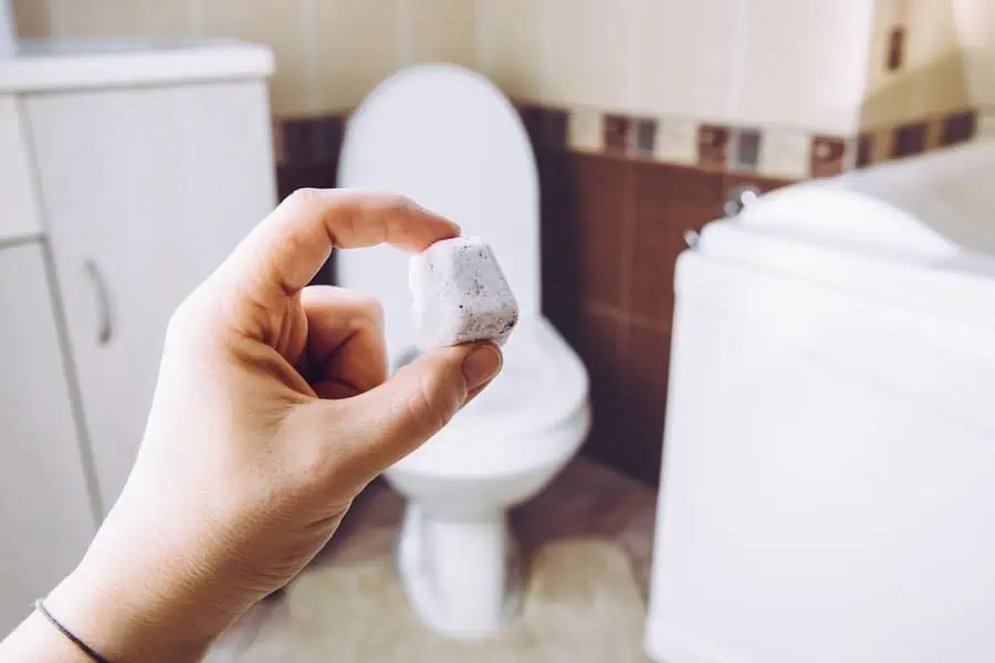 Hand holding a toilet cleaning tablet in front of a toilet in a bathroom