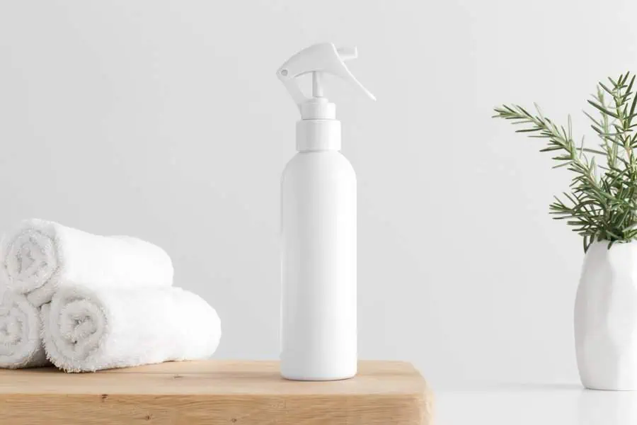 White spray bottle on wooden surface beside rolled towels and plant in vase