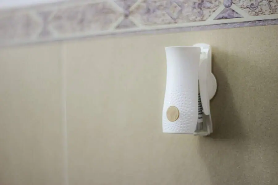 A white automatic air freshener is mounted on a beige tiled wall