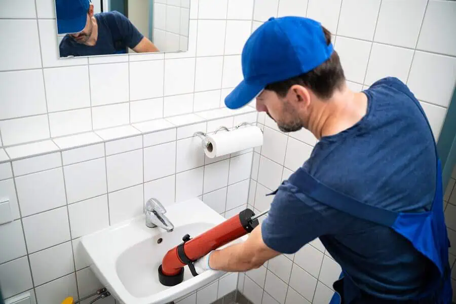 A plumber unclogs a white bathroom sink using a red drain snake